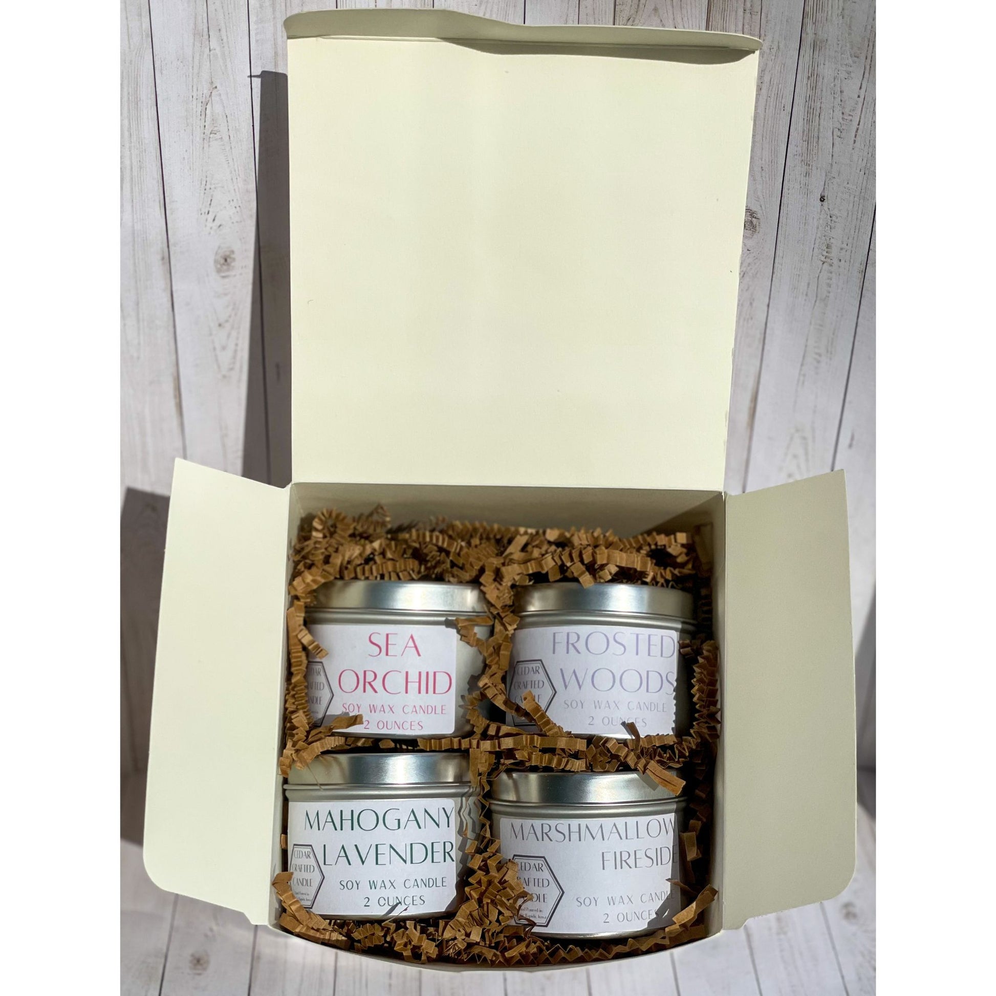 Stack of four silver candle tins, with white labels, in a white box and brown crinkle paper. In the scents of: Sea Orchid, Frosted Woods, Mahogany Lavender, and Marshmallow Fireside